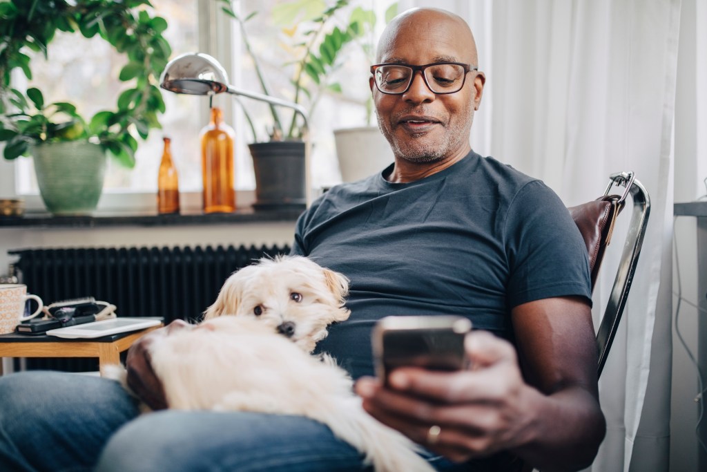 Smiling retired senior male using smart phone while sitting with dog in room at home A Consumer on his cellphone using Digital retailing from Dealer.com