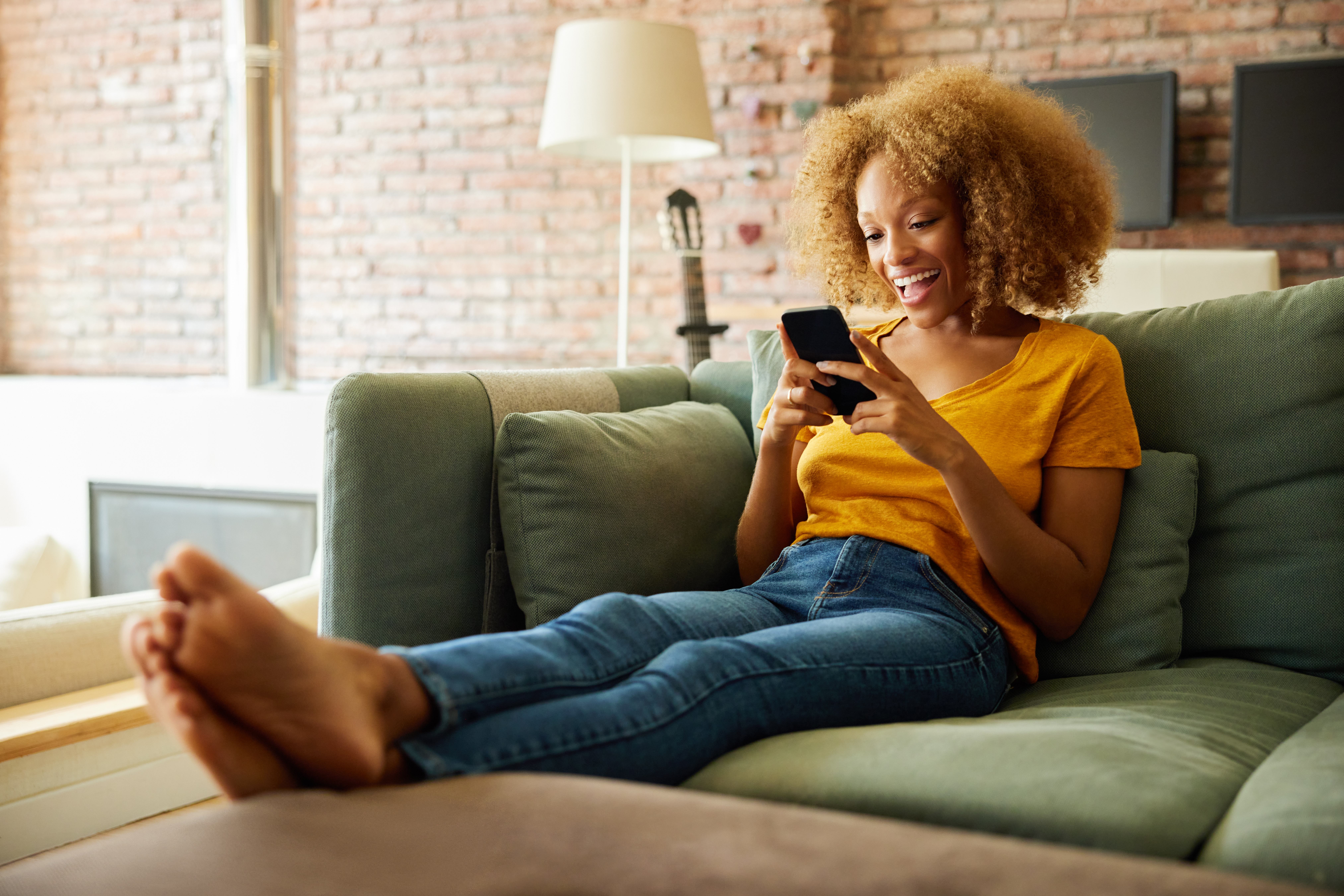 Cheerful woman text messaging through smart phone. Young female with curly hair in living room. She is sitting on sofa at home.